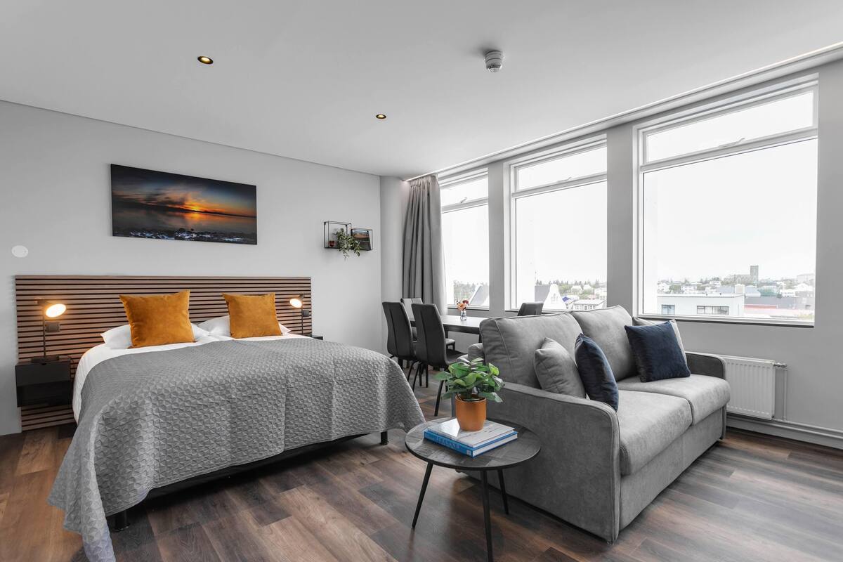 Een moderne hotelkamer met een groot bed, een bank, een klein tafeltje met een plant en uitzicht op de stad door grote ramen.