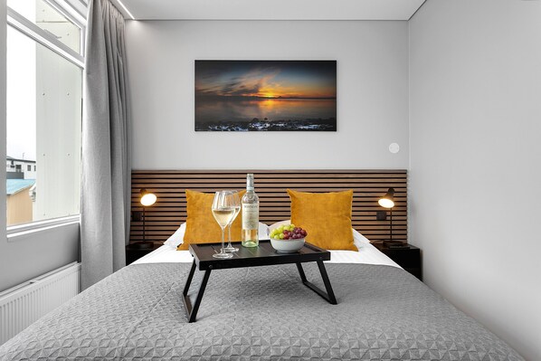 Habitación de hotel con cama, mesita de noche con una botella y vasos, un frutero y un cuadro de atardecer en la pared.