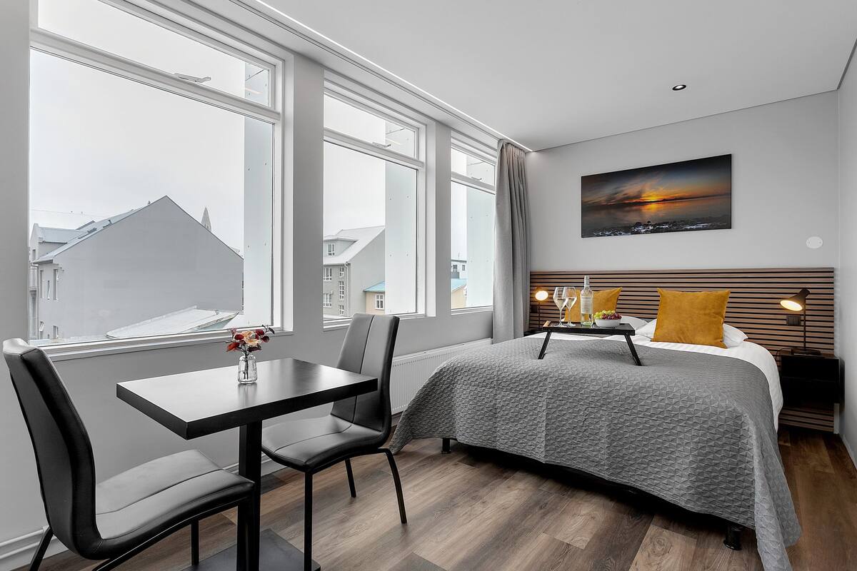 Een moderne hotelkamer met een groot raam, een bed met een bedspreide in geometrische patronen, een nachtkastje met een lamp en een schilderij van een zonsondergang aan de muur.