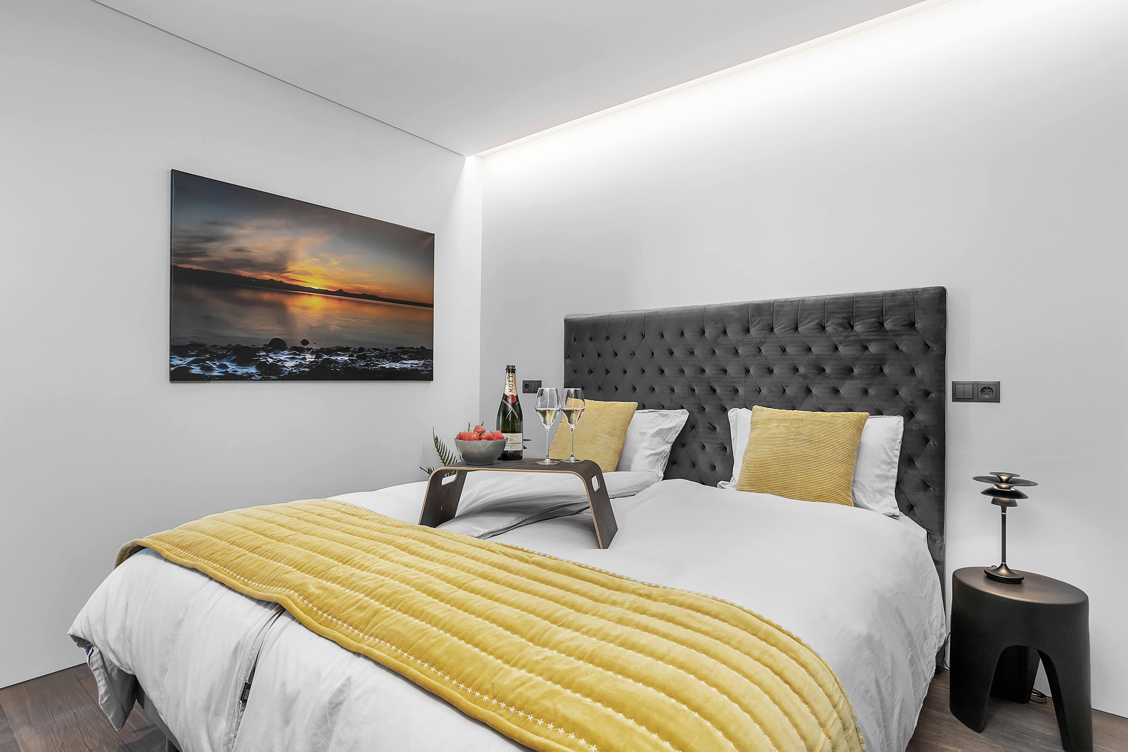 A modern bedroom with a large bed, a bedside table, a nightstand, a wall-mounted lamp, and a sunset painting.