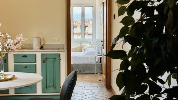 Une pièce comprenant un coin cuisine, une table à manger avec des chaises, et donnant sur une chambre à coucher par une porte ouverte.