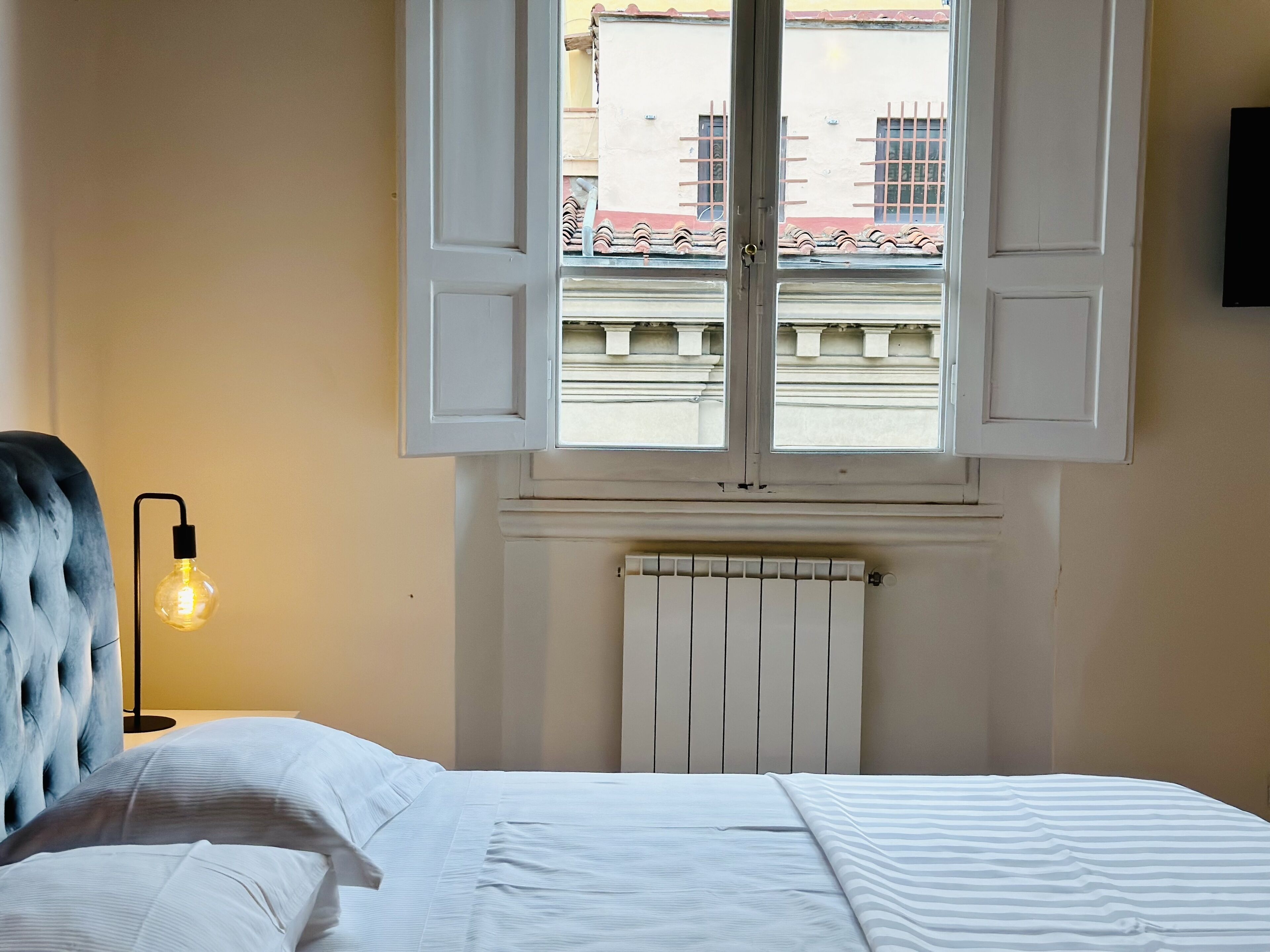 A bedroom with a bed, a radiator, a window with shutters, and a lamp.