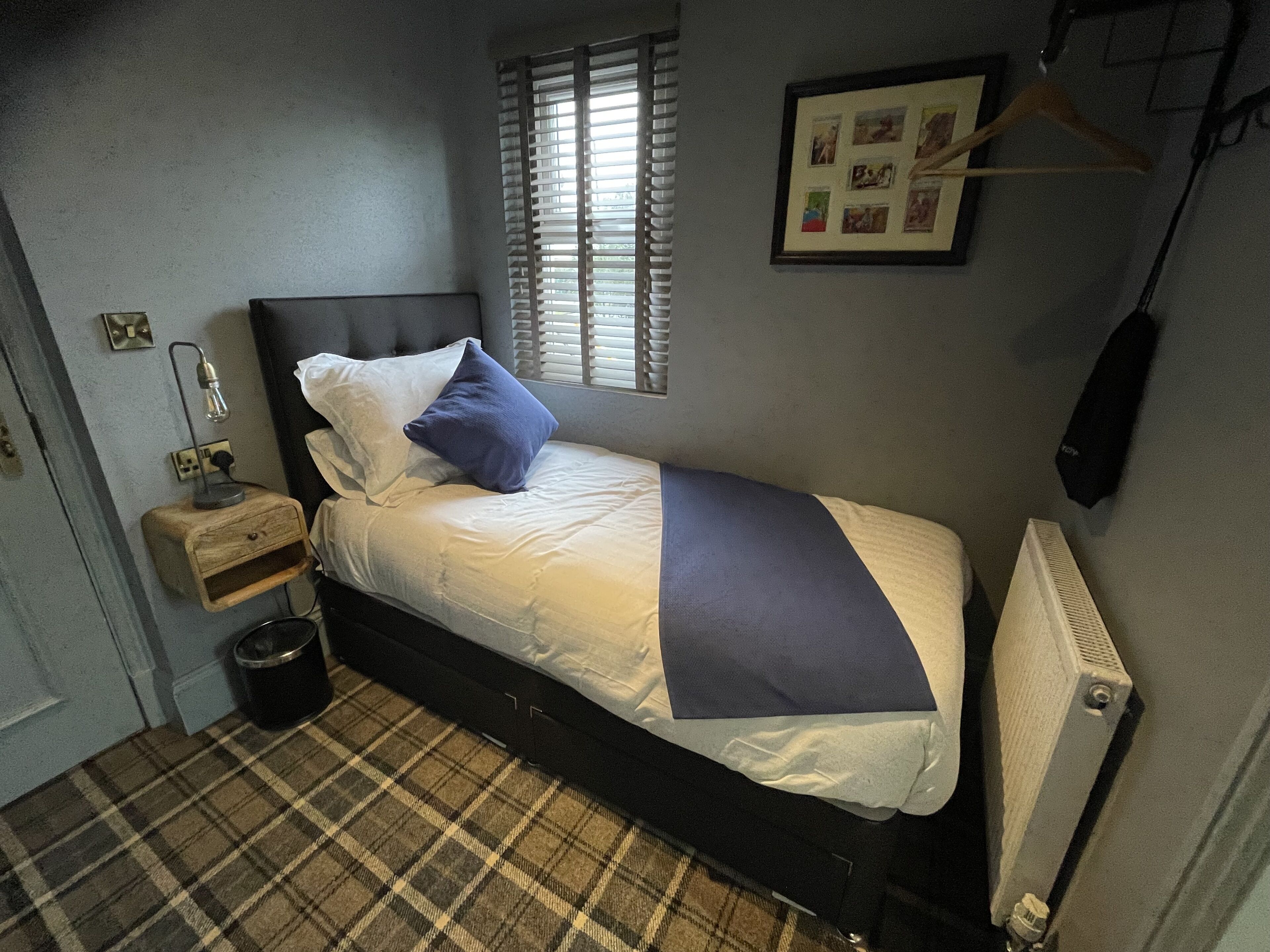 A small, neatly arranged bedroom with a bed, a bedside table, a lamp, a radiator, and a window with blinds.