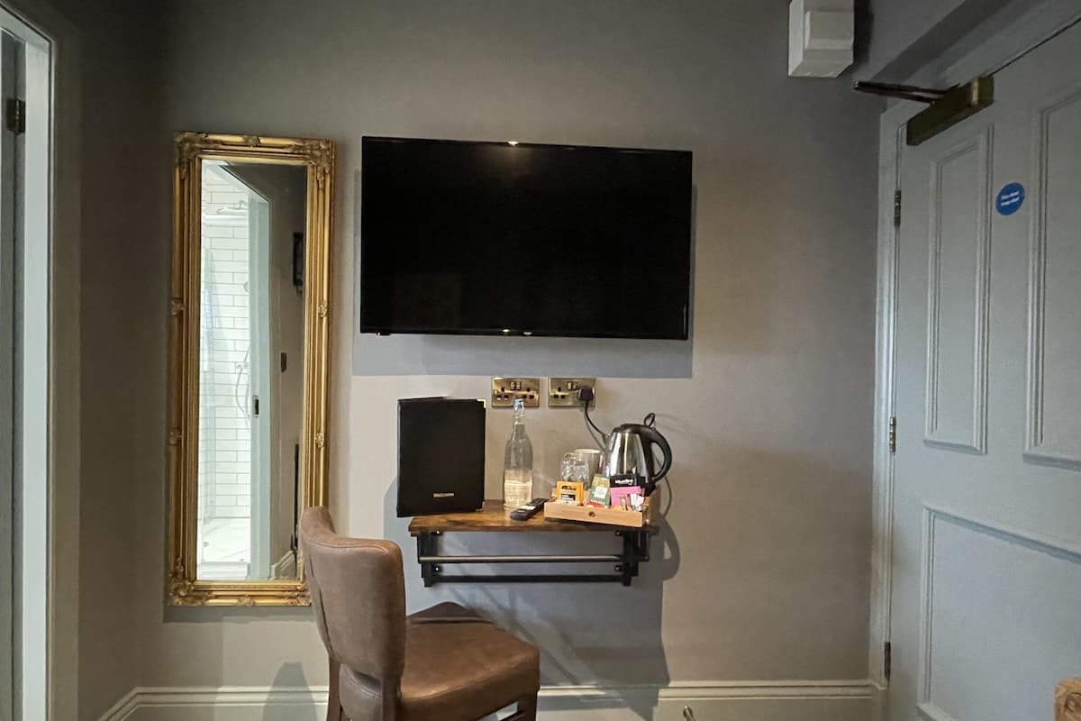 A room with a large mirror, a flat-screen TV mounted on the wall, a wooden desk with a bottle, a kettle, and a chair.