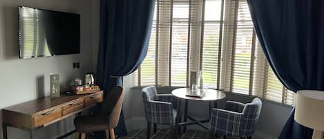 Quarto de hotel com cama, escrivaninha, cadeira, mesa com garrafa e copo, e televisão na parede.