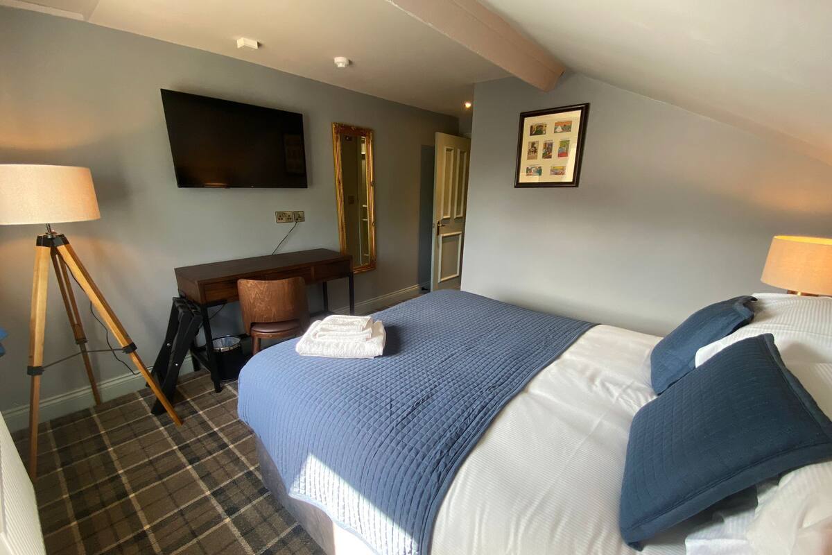 A hotel room with a bed, a desk, a chair, a lamp, a mirror, and a framed picture on the wall.