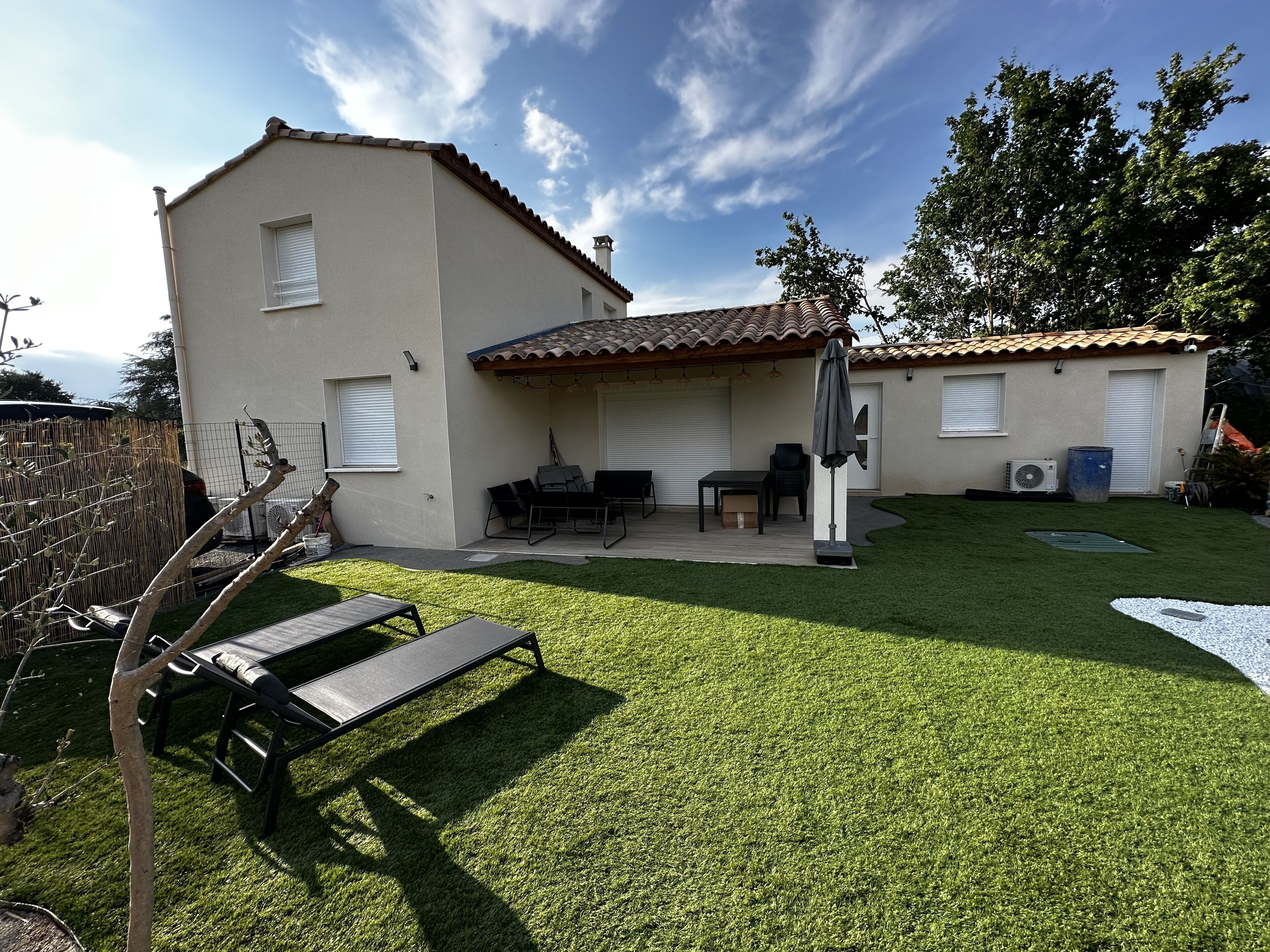 Ein zweistöckiges Haus mit Terrasse, Sitzgelegenheiten im Freien und einem kleinen Garten.