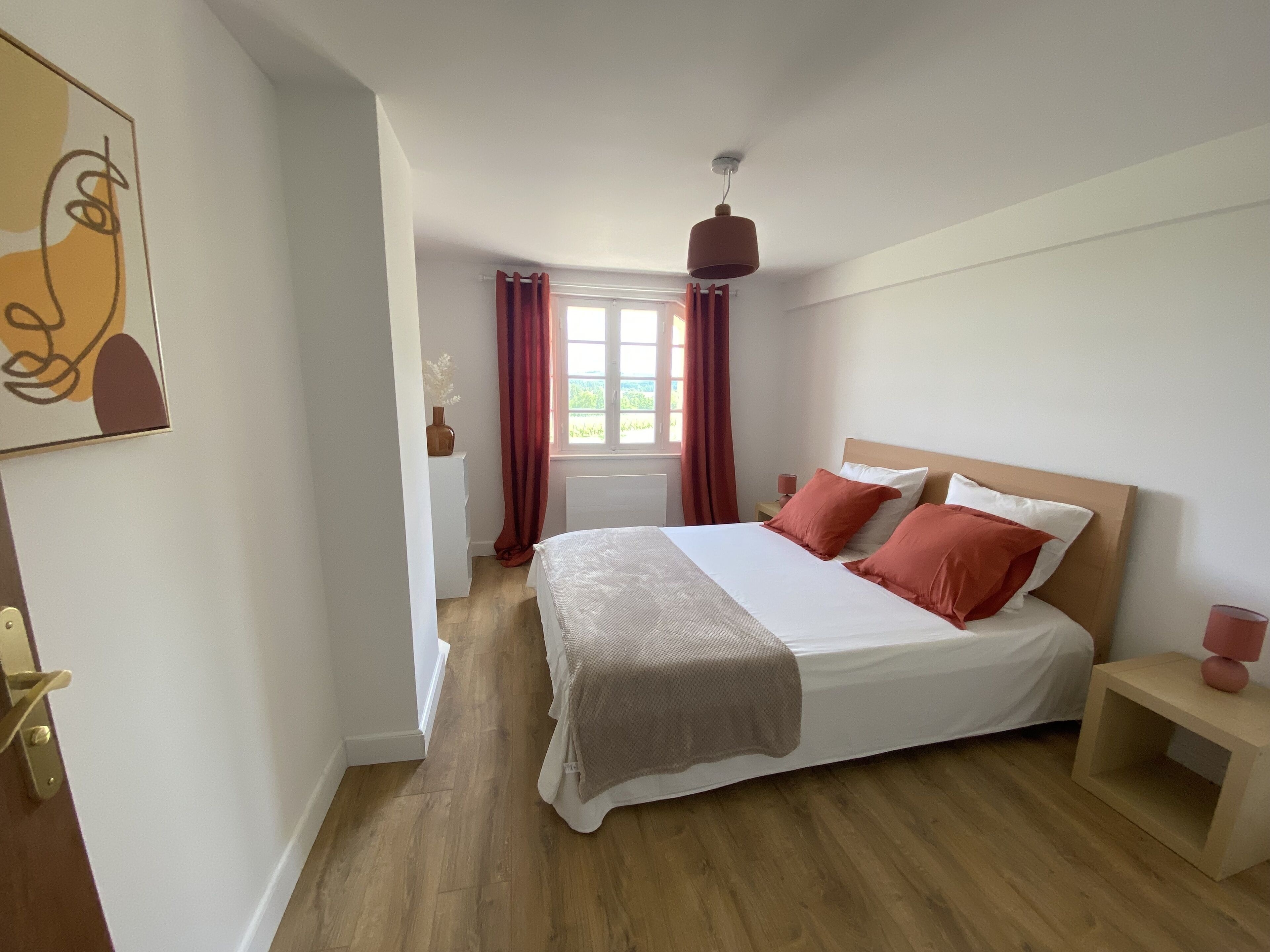 Un dormitorio con una cama grande, una mesita de noche, una ventana con cortinas y una lámpara colgante.