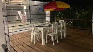 An outdoor dining area with white tables and chairs, a colorful umbrella, and a glass enclosure with a view of a pool and people. - "Beach Haven Nilaveli" a home away from home (Nilaveli, Eastern Province, Sri Lanka)