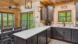 A kitchen with dark wooden cabinets, granite countertops, and a built-in dishwasher. There are two windows with blinds, a ceiling fan, and a sign welcoming guests. - *New Build* 3 BR w/ Hot Tub, Arcade, WiFi, Catch and Release Pond, Free Tickets (Sevierville)