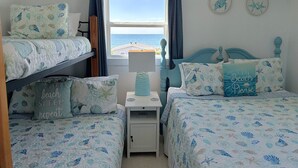 Una camera da letto con un letto a castello, un letto singolo e un comodino con una lampada. Si vede una finestra con tende, vista mare e due decorazioni a parete.