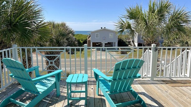 Un terrazzo in legno con due poltrone Adirondack color turchese e un tavolino, con vista su una spiaggia e una casa bianca con veranda.