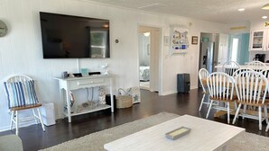 Un soggiorno moderno con una TV a schermo piatto, arredi bianchi e un'area pranzo con sedie in legno.