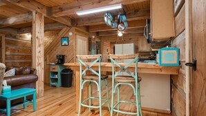 Un intérieur de chalet chaleureux, avec un revêtement mural en bois, une kitchenette, un coin repas doté de tabourets de bar et un espace de vie comprenant un canapé et une table basse.