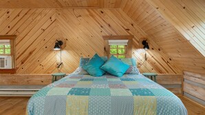 Une chambre mansardée chaleureuse avec un plafond en pente en bois, une couvre-lit matelassé, deux lampes de chevet et une fenêtre donnant sur un paysage verdoyant.
