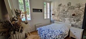 Een slaapkamer met een bed, een nachtkastje, een wandheater, een raam met uitzicht op groen en een muurschildering van tropische planten.
