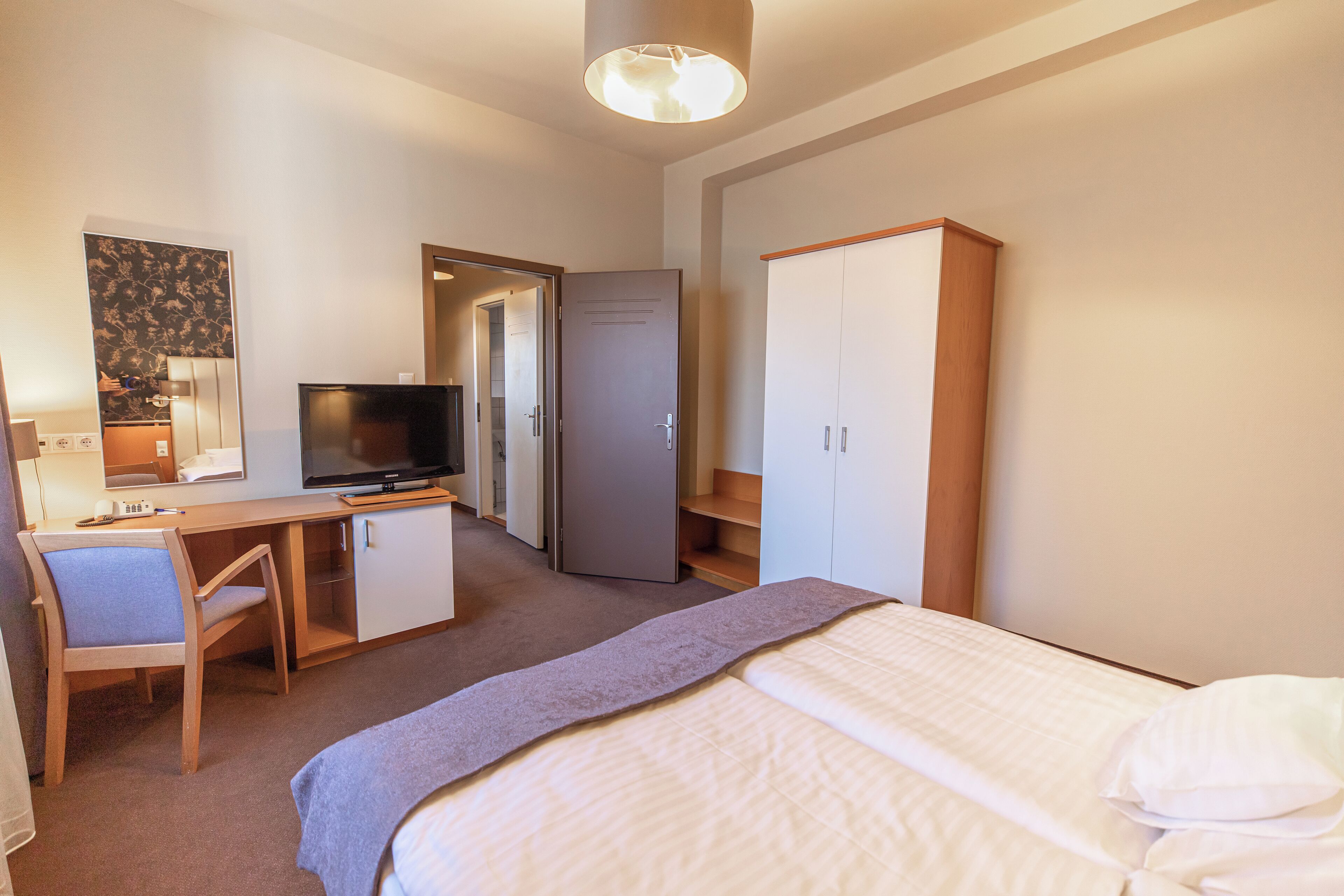 A hotel room with a bed, a desk with a chair, a television, a wardrobe, and a door leading to a bathroom.