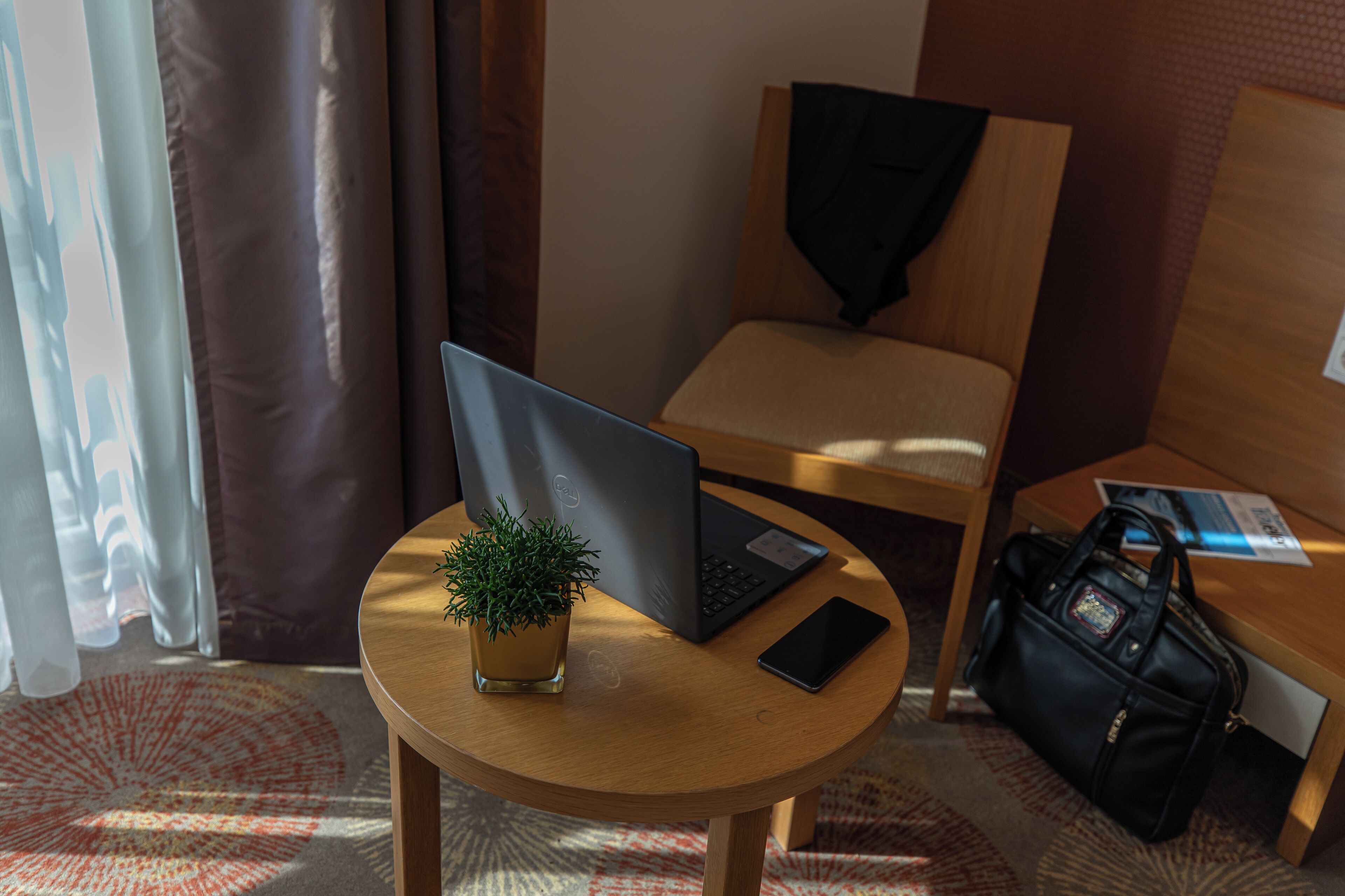 Habitación de hotel con un escritorio de madera, una computadora portátil, un teléfono inteligente, una pequeña planta en maceta y un bolso negro en el suelo.