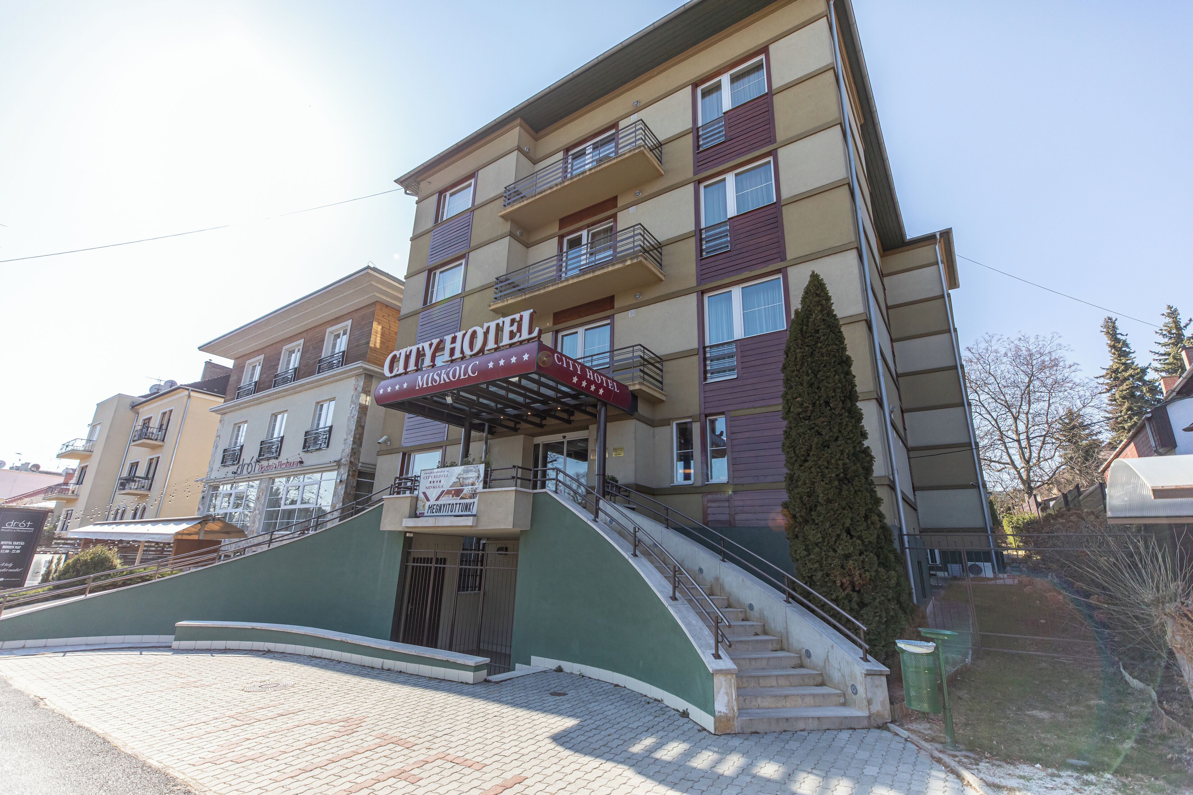 Un hotel de varios pisos con un letrero llamativo que dice “CITY HOTEL”. Tiene una escalera que conduce a la entrada, un toldo verde y una vista despejada del entorno.