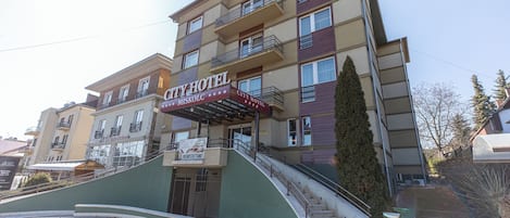 Un hotel de varios pisos con un letrero llamativo que dice “CITY HOTEL”. Tiene una escalera que conduce a la entrada, un toldo verde y una vista despejada del entorno.