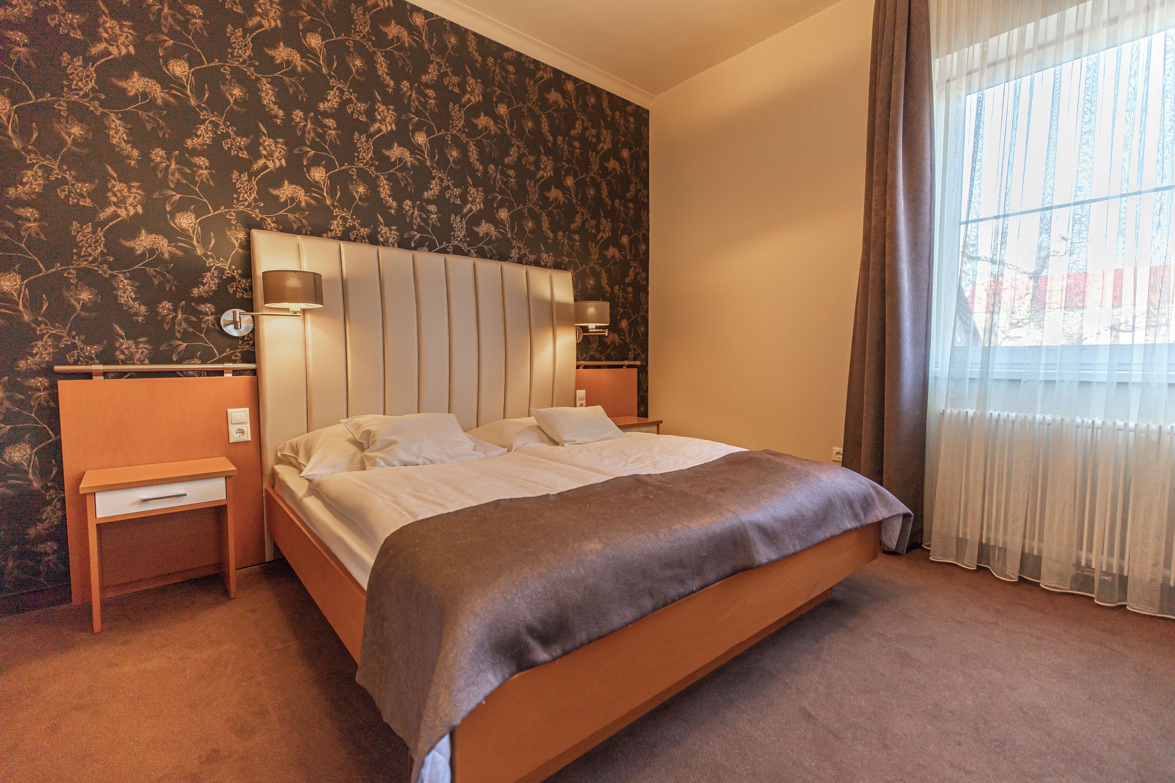A hotel room with a large bed, two bedside tables, a nightstand, a wall-mounted lamp, and a window with curtains.