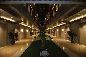 A long, well-lit corridor with a central green carpet, flanked by potted plants and illuminated wall sconces. - Shree Jee Vilas (Sonepat)