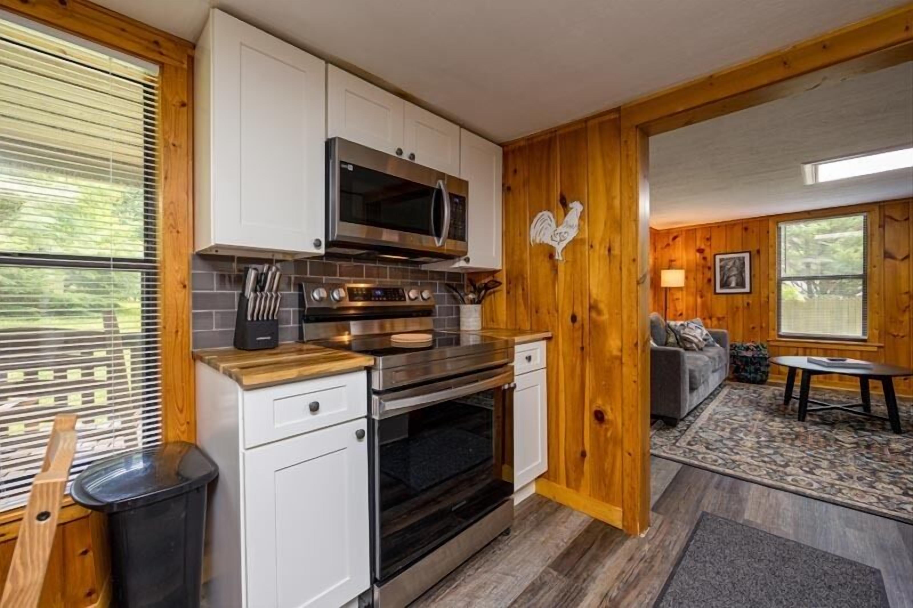 Une cuisine compacte avec des meubles blancs, un four à micro-ondes en acier inoxydable et un four traditionnel. Un mur en bois est décoré d’un motif de coq, et une fenêtre est dotée de stores.