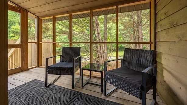 A covered porch with two wicker chairs and a small table, wooden railings, and a view of a wooded area.