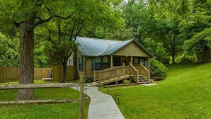 A small, single-story cabin with a porch and a metal roof, surrounded by lush greenery and a wooden fence.