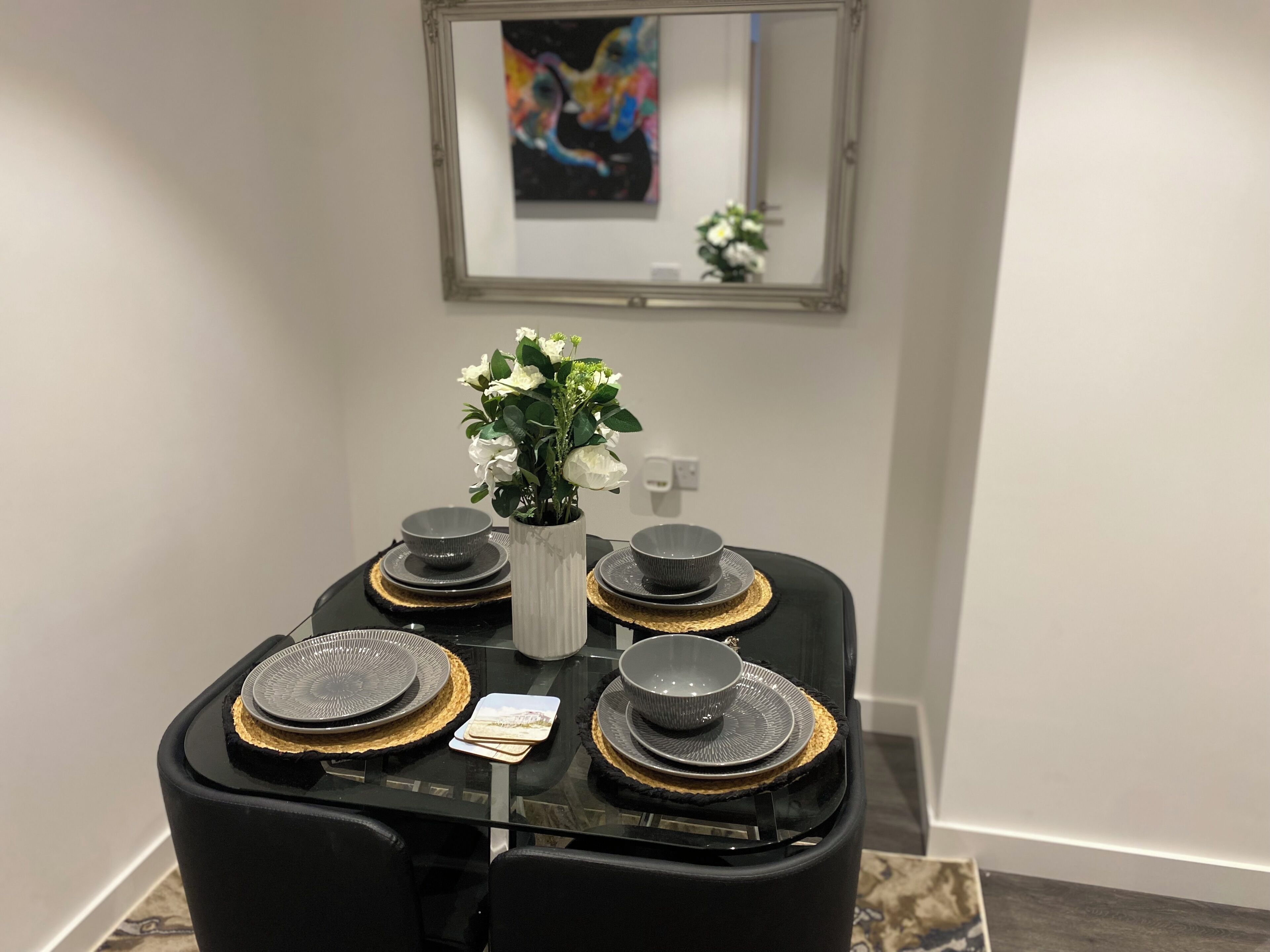 Un comedor con una mesa dispuesta para cuatro personas, destacando platos negros con detalles dorados, un florero blanco con flores blancas y un cuadro abstracto enmarcado en la pared.