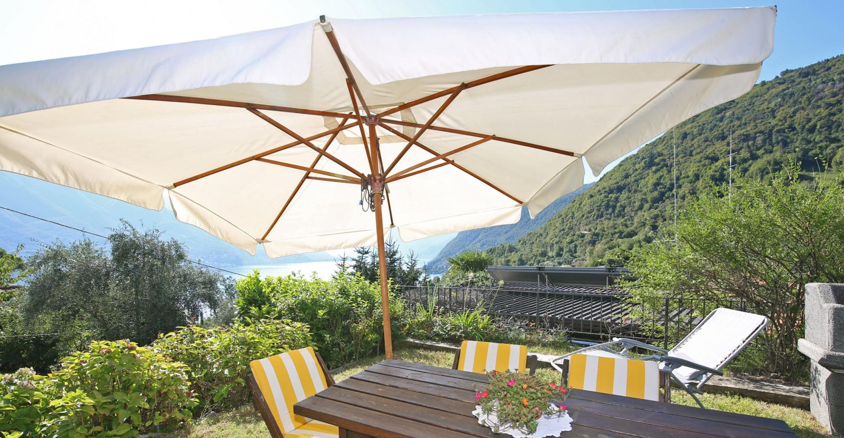 An outdoor seating area with a wooden table and chairs, a large white umbrella, and a mountainous backdrop.