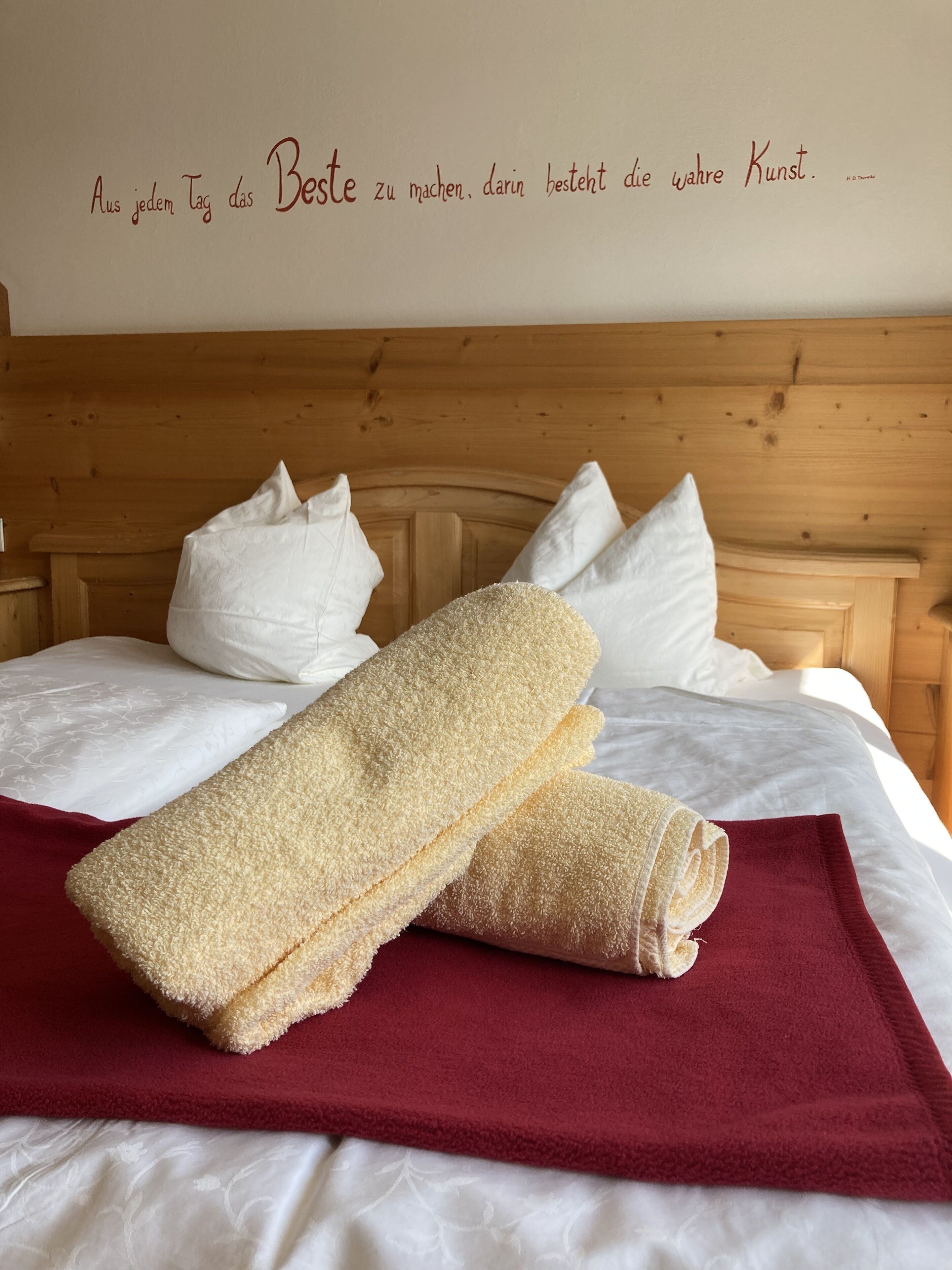 A neatly made bed with white linens, a red blanket, and two beige towels.