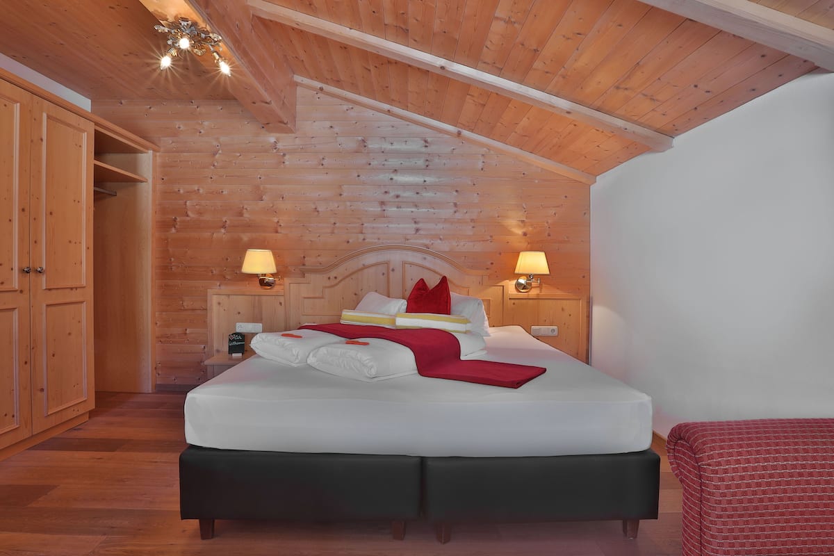 A bedroom with a wooden ceiling, a bed with white bedding and red accents, two bedside lamps, and a wooden wardrobe.