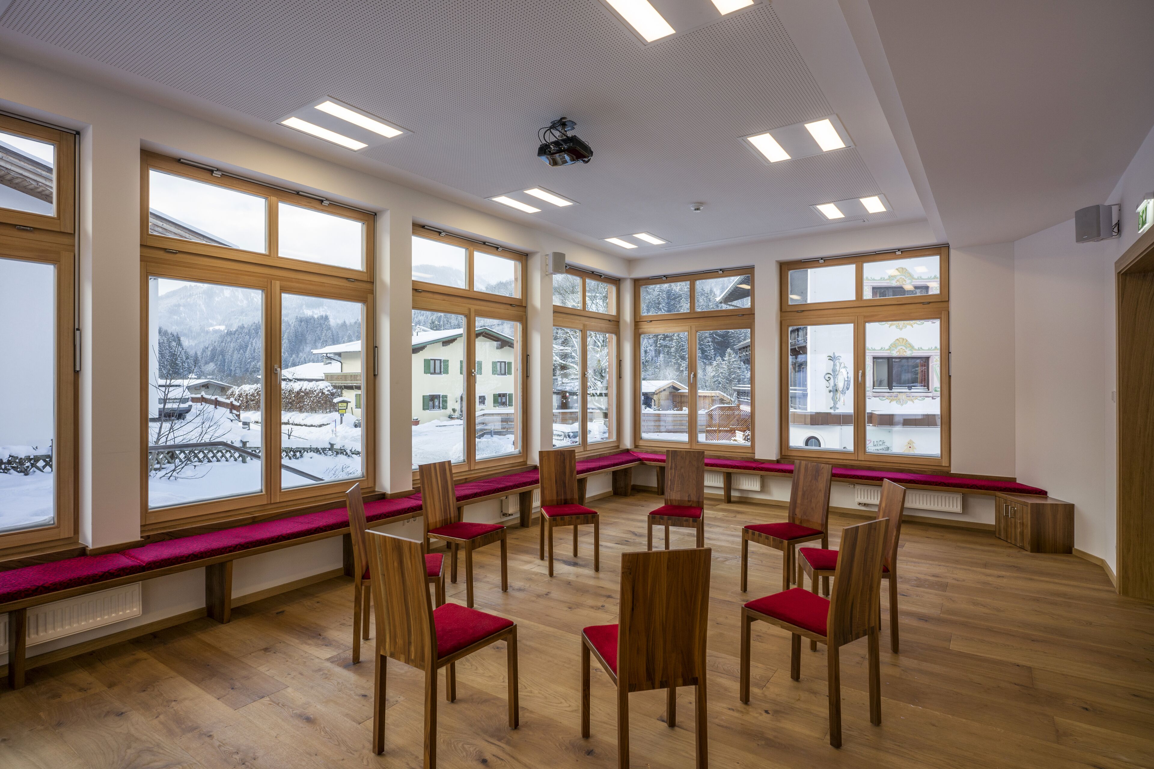 A room with large windows, wooden chairs with red cushions, and a wooden floor.