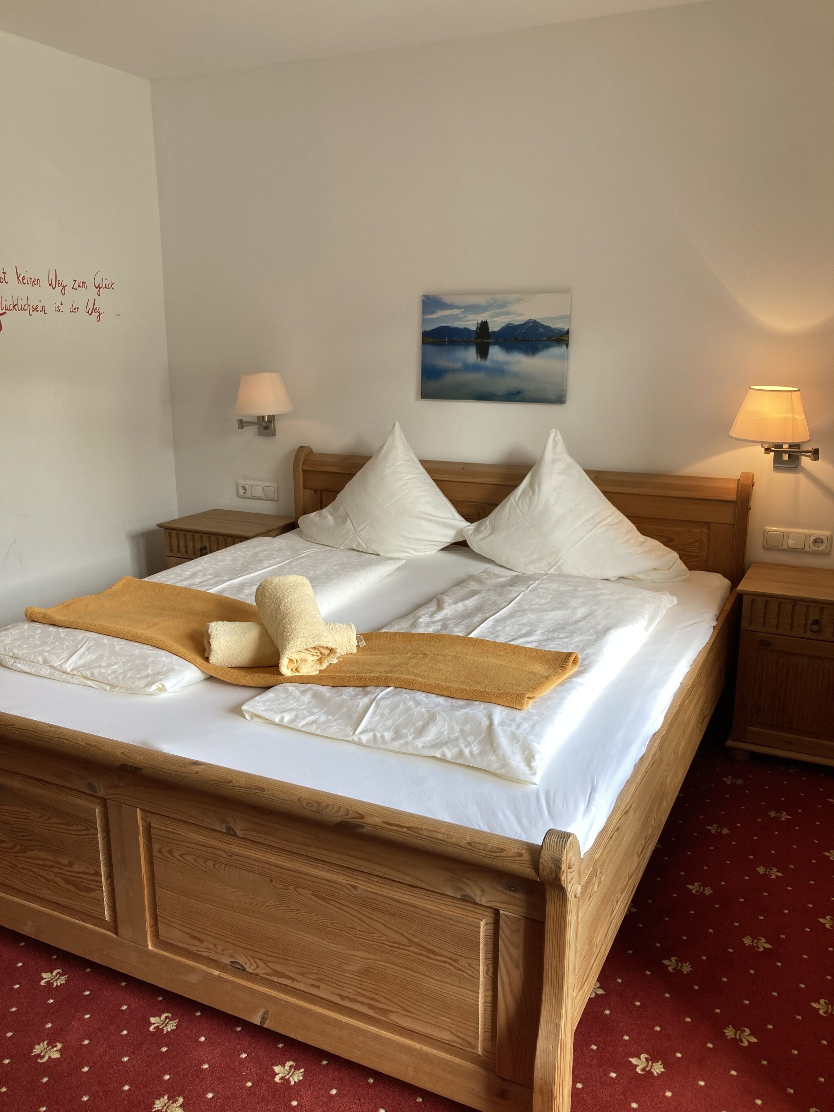 A neatly made bed with white linens and a brown throw blanket, wooden headboard, two bedside tables with lamps, a wall-mounted painting of a lake, and a red carpet with a floral pattern.