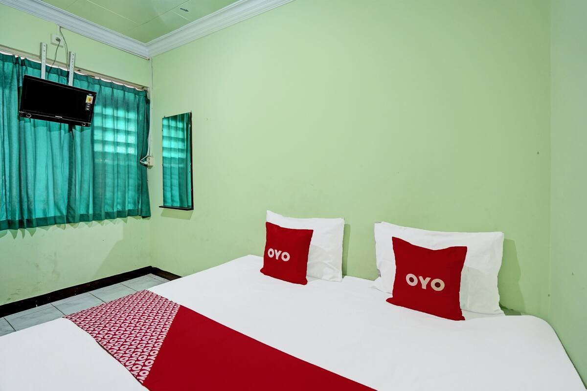 Una habitación de hotel con cama, dos almohadillas rojas con la palabra "OYO" y un televisor montado en la pared, además de una ventana con cortinas.