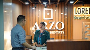 A reception desk with a wooden panel background, a sign for "ARZO GROUP OF CONSUMERS RESORTS", and a name tag on the staff member. - ARZO Hotel Premier (Makati)