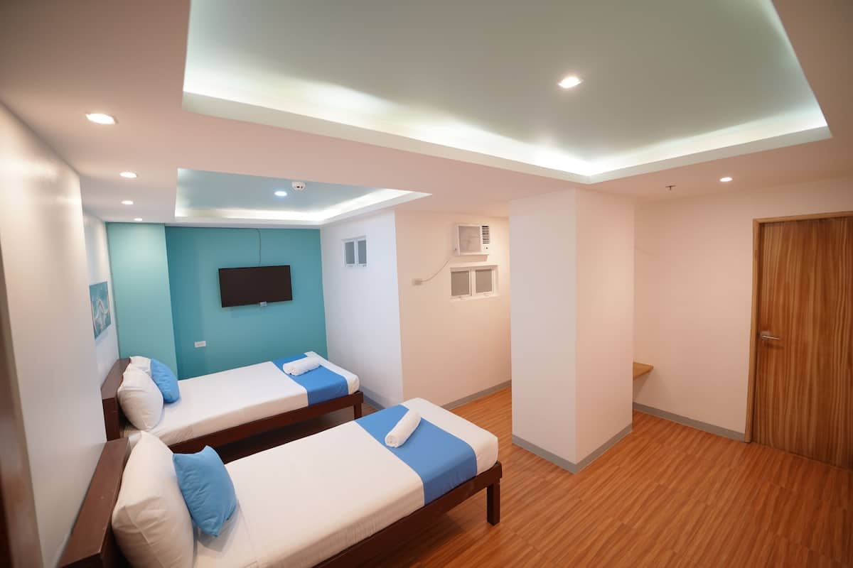 Ein Hotelzimmer mit zwei Einzelbetten, einem flachbildfernseher an der Wand, einem Deckenventilator und einer Holztür.