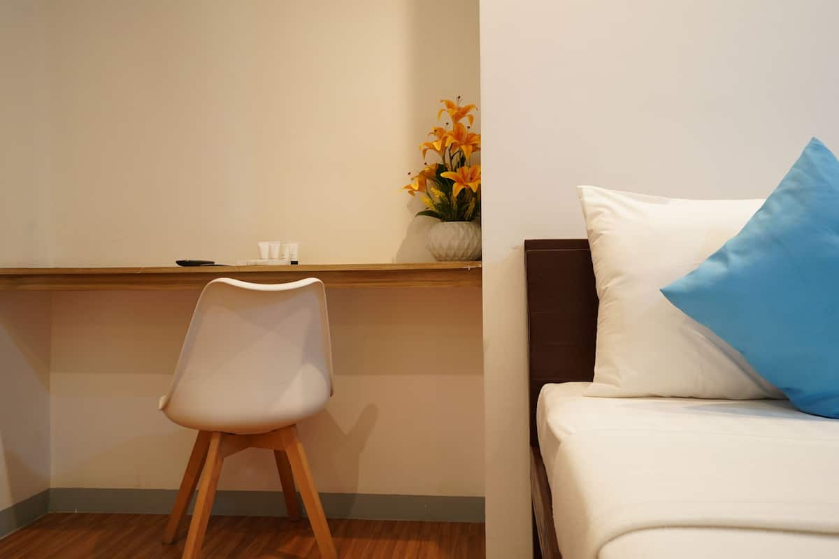 Ein Hotelzimmer mit einem Holztisch, einem weißen Stuhl, einem Bett mit blauem Kopfkissen und einer Wandvase mit Blumen.