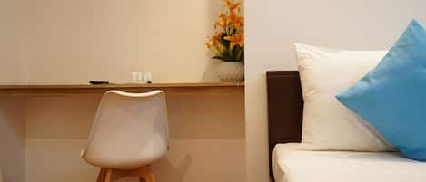 A hotel room with a wooden desk, a white chair, a bed with a blue pillow, and a vase with flowers on the wall.