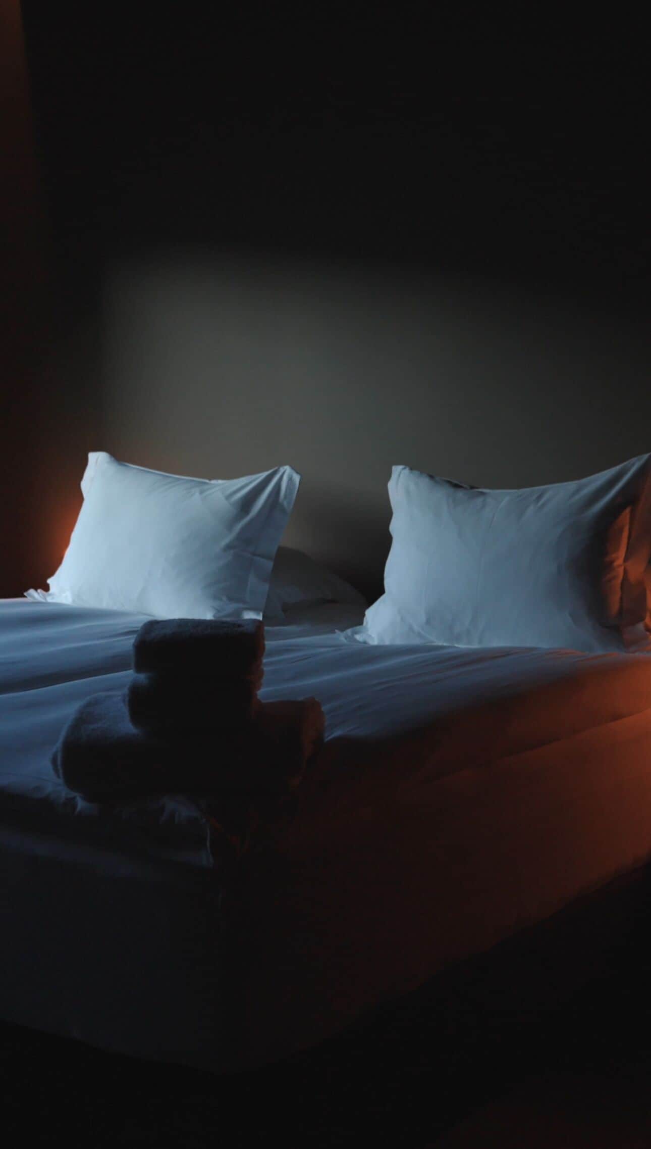 Quarto de hotel pouco iluminado com uma cama, almofadas e um ursinho de pelúcia na cama.