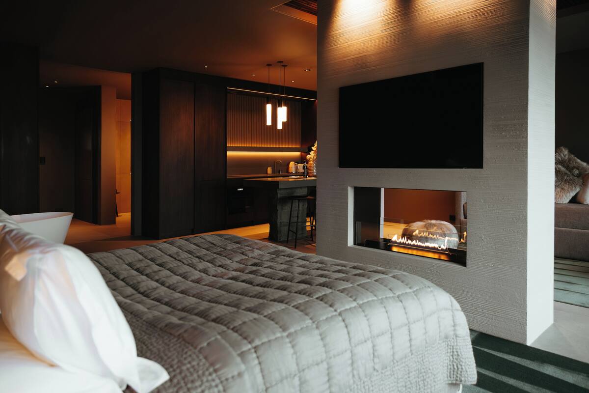 A modern hotel room with a large bed, a flat-screen TV mounted on the wall, a small kitchen area, and a fireplace.
