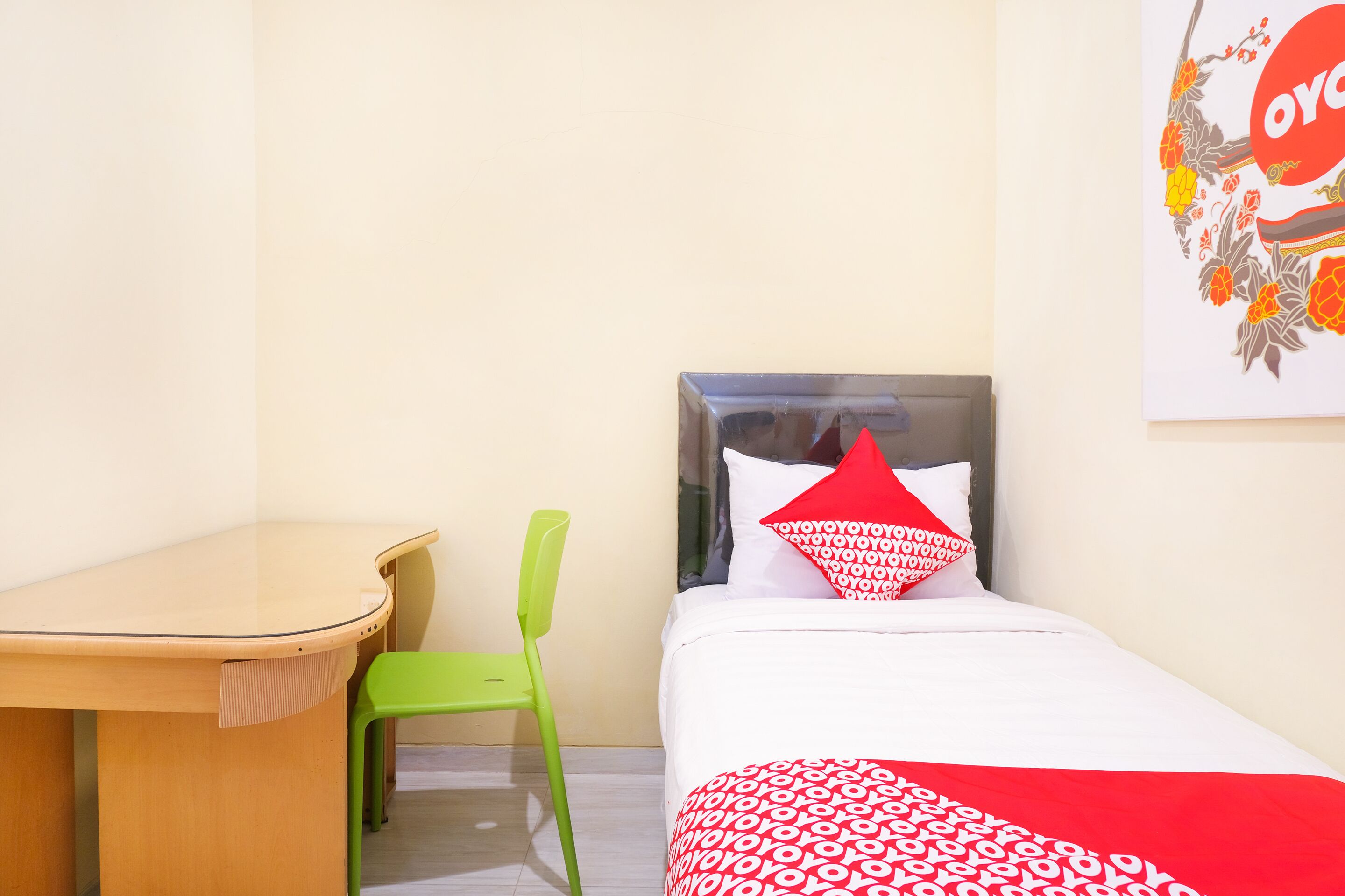 A small room with a single bed, a green chair, a wooden desk, and a wall-mounted artwork.
