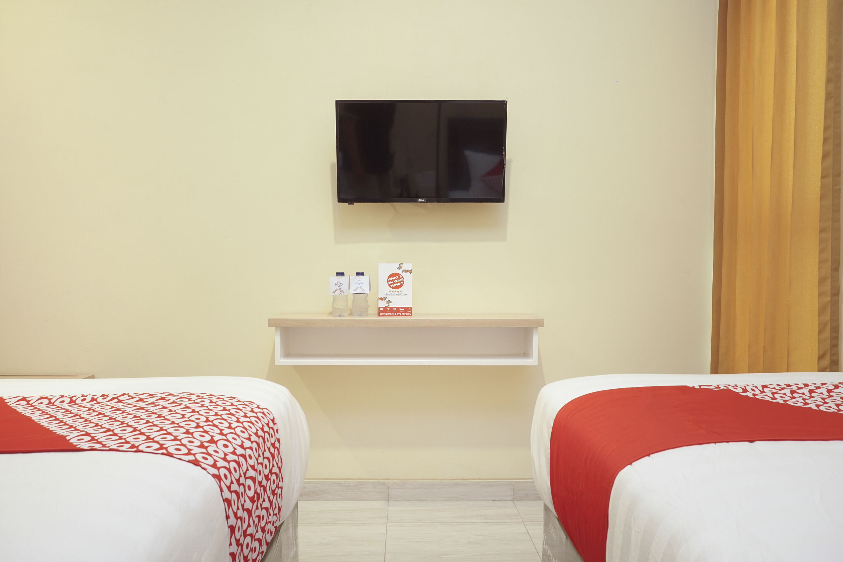 A hotel room with two beds, a TV mounted on the wall, a shelf with toiletries, and a curtain on the window.