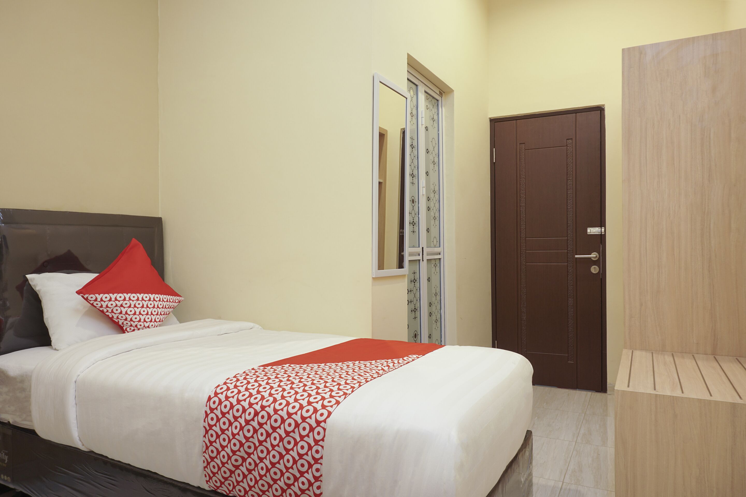 A neatly made bed with a patterned bedspread and pillows, a wooden wardrobe, and a door with a mirror.
