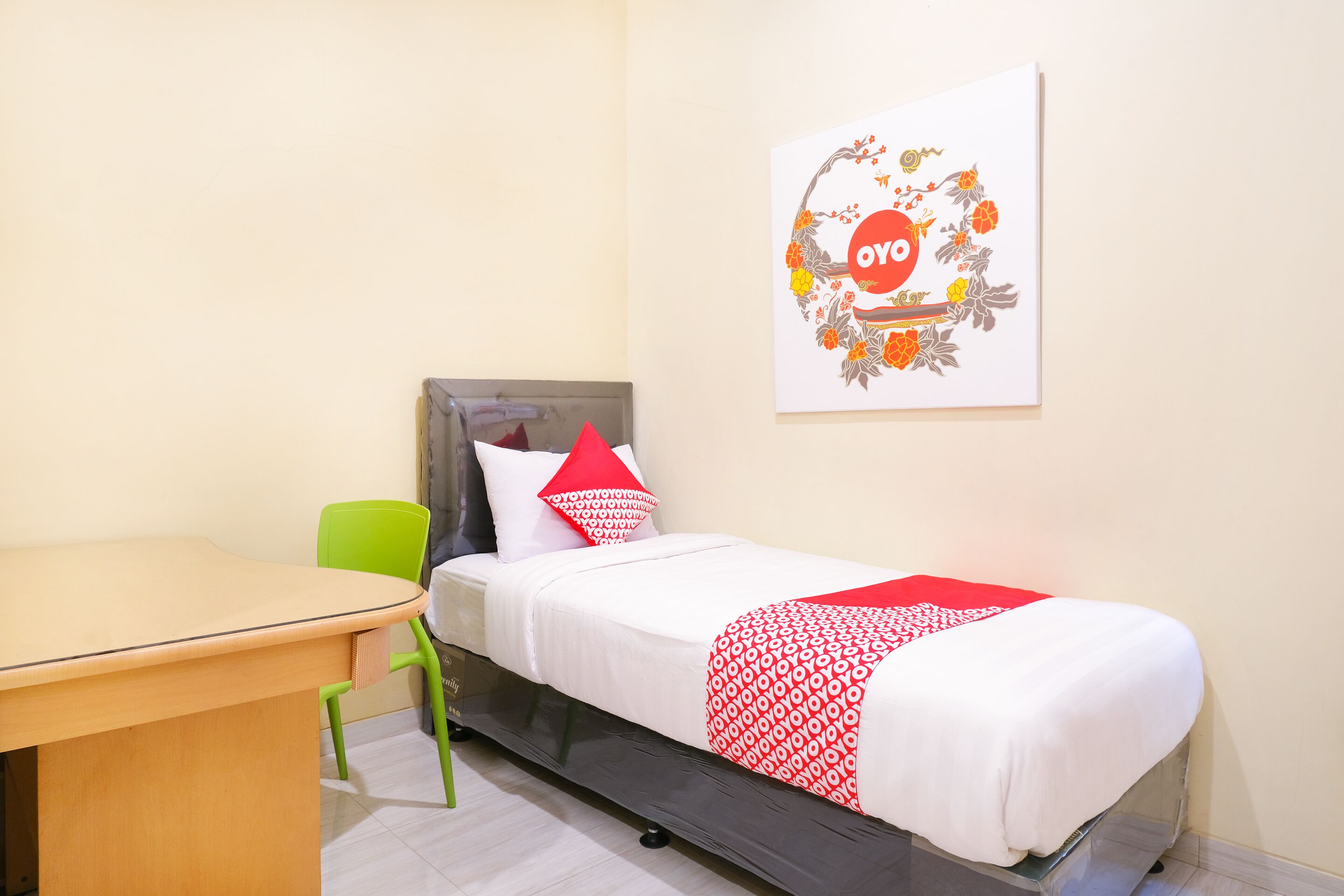 Un lit simple avec une couvre-lit à motifs rouges et blancs, une chaise verte, une table en bois et une œuvre d’art fixée au mur, présentant un motif circulaire.