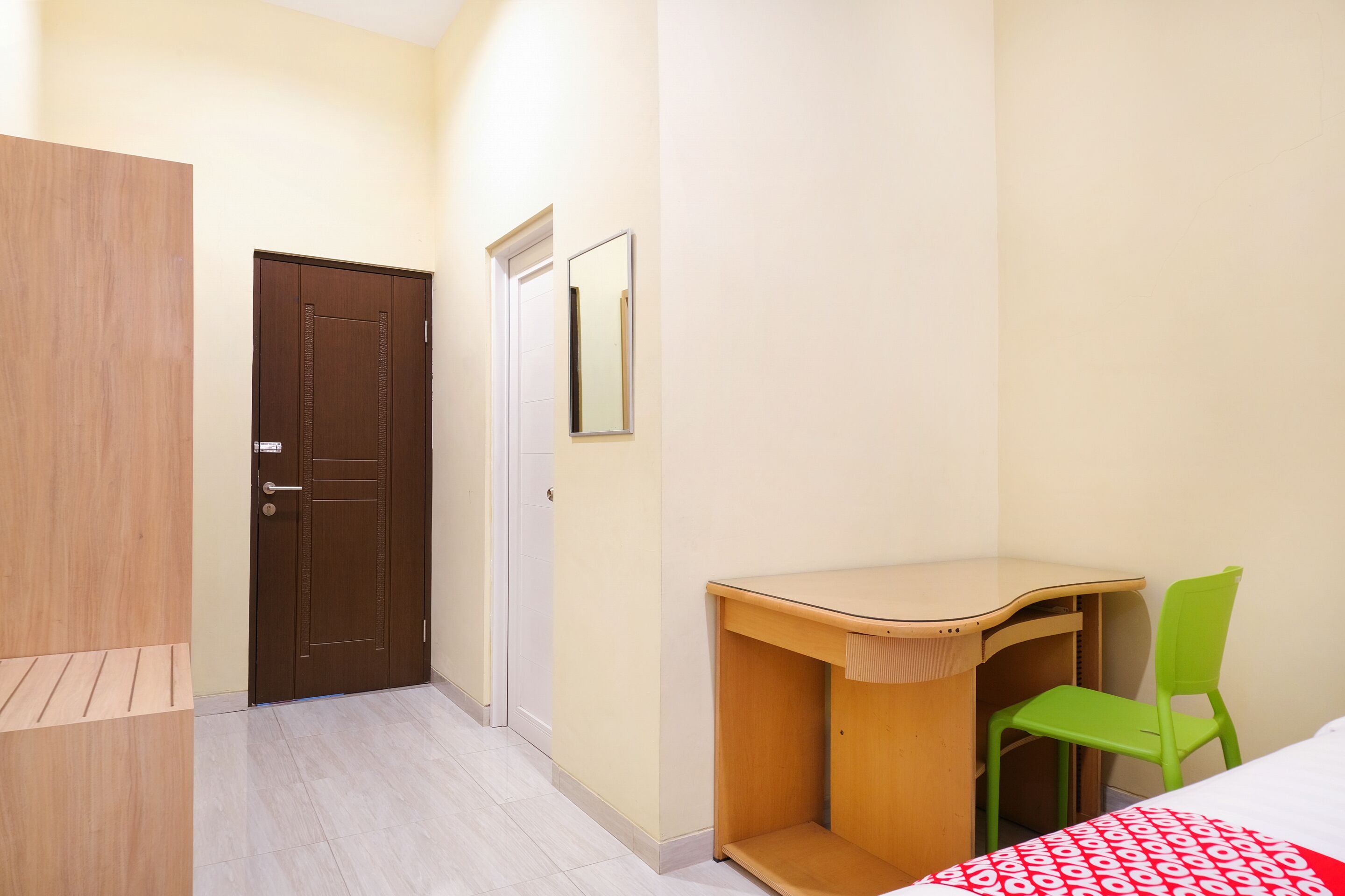 A room with a wooden wardrobe, a door, a mirror, a desk with a chair, and a bed with a patterned bedspread.