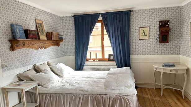 Une chambre à coucher avec un lit, une petite table, une étagère remplie de livres et une fenêtre avec des rideaux.