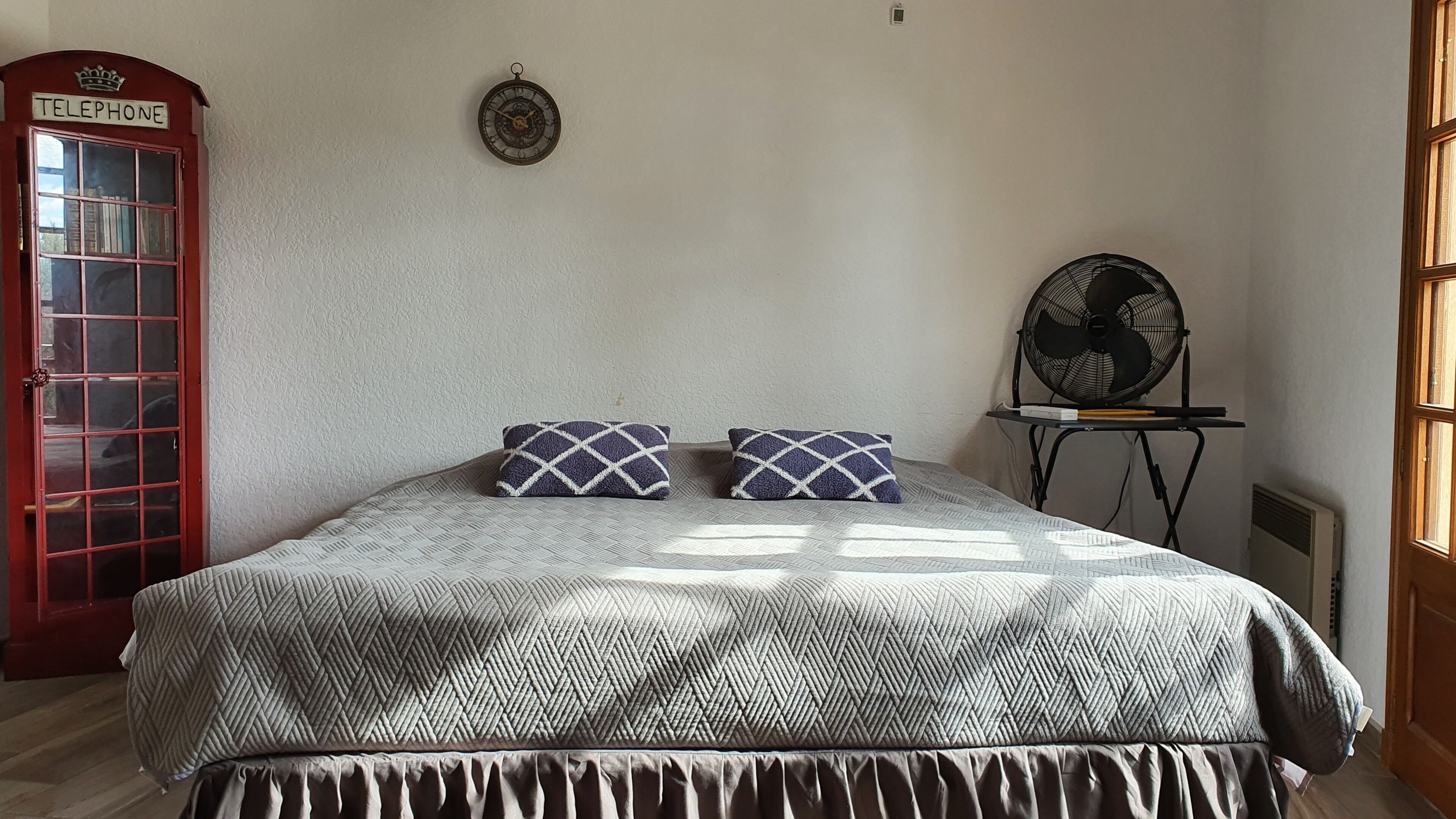 Een slaapkamer met een bed, twee gestreepte kussens, een wandklok, een ventilator, een radiator en een rode telefooncel.