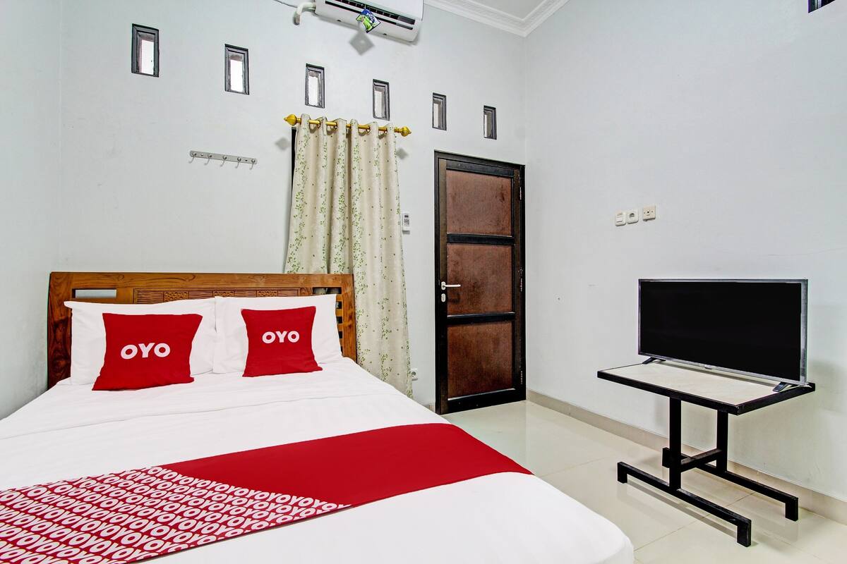 A neatly made bed with red and white bedding, a television on a stand, and a door with a window.